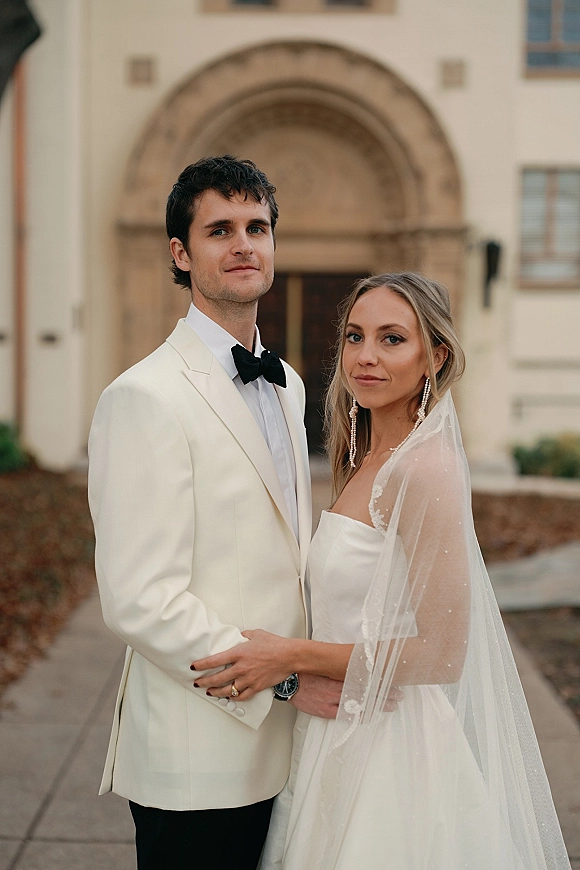 Couple portrait of bride in strapless wedding dress and veil with drop earrings, and groom in white dinner jacket at arched doorway entrance