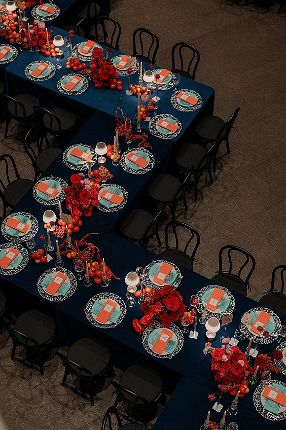 Reception tablescape with a navy wedding table, red floral centerpieces, taper candles, teal napkins, patterned plates, and produce on a long banquet table in a hall
