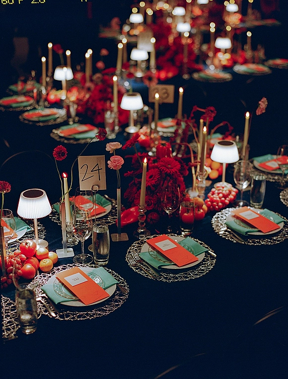 Reception tablescape with candlelit wedding table glow, red floral centerpieces, citrus, teal napkins, and layered place settings in a dim room