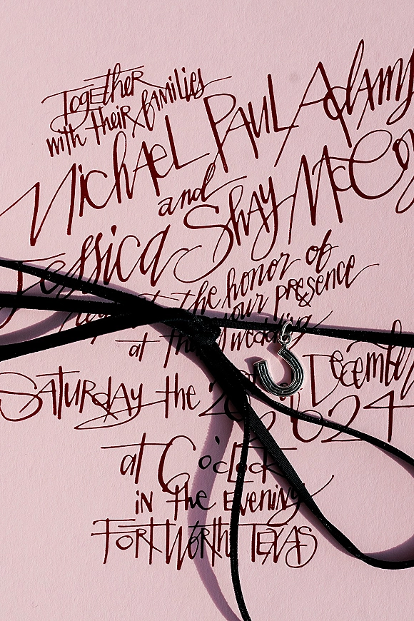 Wedding invitation with calligraphy lettering, tied with a black ribbon and horseshoe charm on soft pink paper background