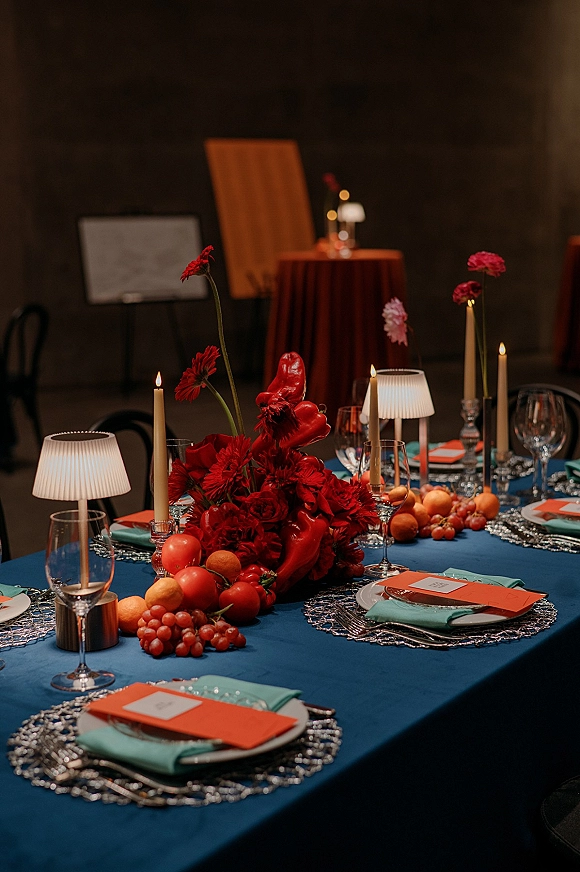 Reception tablescape with a red floral centerpiece, taper candles, mini lamps, crystal glassware, orange place cards on a blue tablecloth
