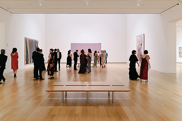 Wedding reception guests mingling at wedding cocktail hour in formal attire, chatting by wall art and a wooden bench in a white museum gallery