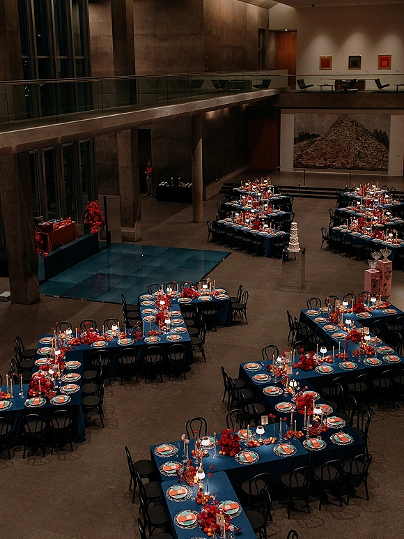 Reception tablescape with a banquet table layout featuring blue tablecloths, red floral centerpieces, taper candles and lamps in a modern atrium venue