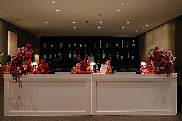 Wedding bar setup with wedding bar decor featuring red and pink florals, fruit display, menu cards, glassware, and black back bar shelves