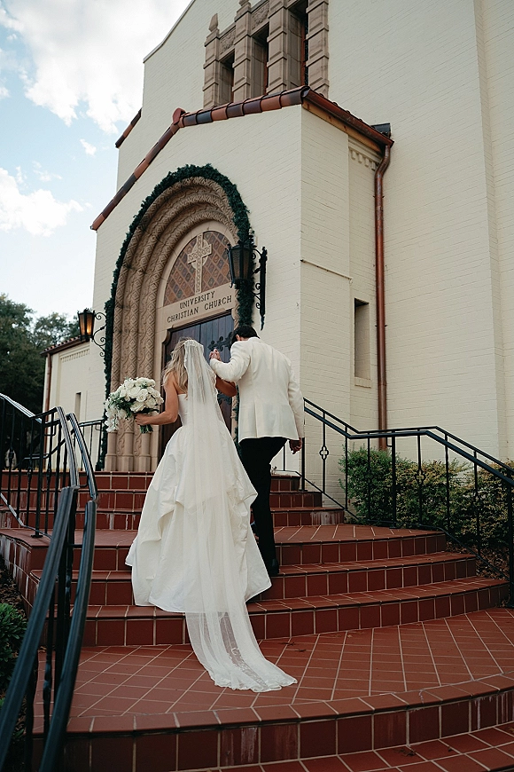 Wedding ceremony exit as newlyweds walk up brick church steps, bride’s long veil and train trailing, holding a white bouquet under arched doorway