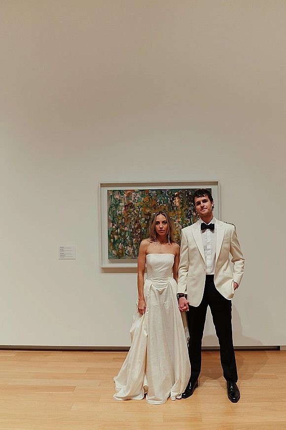 Couple portrait of bride and groom holding hands, she in a strapless gown and he in a white tux, beside framed art on a white gallery wall