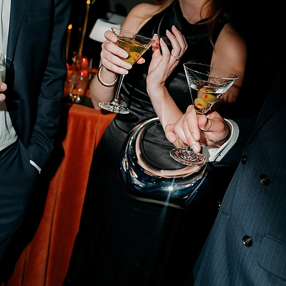 Wedding cocktails in martini glasses with green olives held over a candlelit bar counter, one hand showing a wedding ring and bracelet