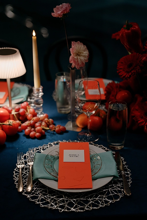 Reception tablescape with a blue tablecloth, clear glass charger, aqua napkin, pink blooms, fruit, and a single taper candle in a dark room