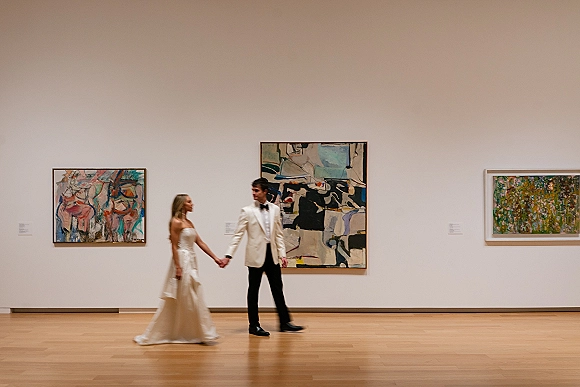 Couple portrait of bride and groom holding hands, her strapless wedding dress and his bow tie in a bright art gallery with framed artwork