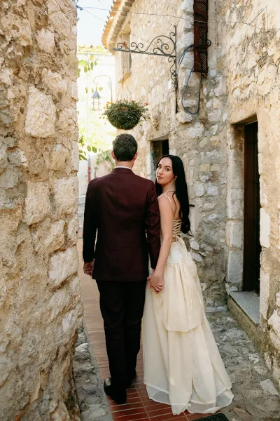 Couple portrait of bride and groom walking away hand in hand, bride looking back in a corset-back gown down a stone alleyway.
