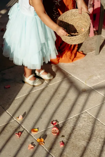 Flower girl in a blue tulle flower girl dress holding a woven basket of rose petals on a sunlit stone tile aisle