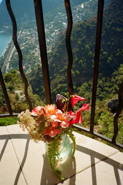 Bridal bouquet with tropical flowers and cascading greenery in a glass vase on a stone ledge, overlooking a mountain valley lake view