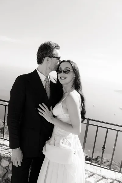 Couple portrait in black and white wedding portrait style, groom in sunglasses kissing bride’s forehead on a stone terrace by the ocean.