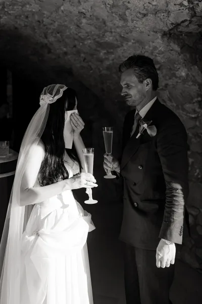 Wedding couple portrait of bride wiping tears as they toast with champagne flutes, veil and rings visible against a shadowed stone wall backdrop