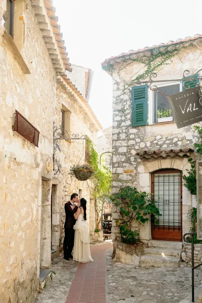 Wedding kiss portrait of the bride and groom kissing on a cobblestone stone alley, framed by an arched doorway and hanging flower basket in daylight