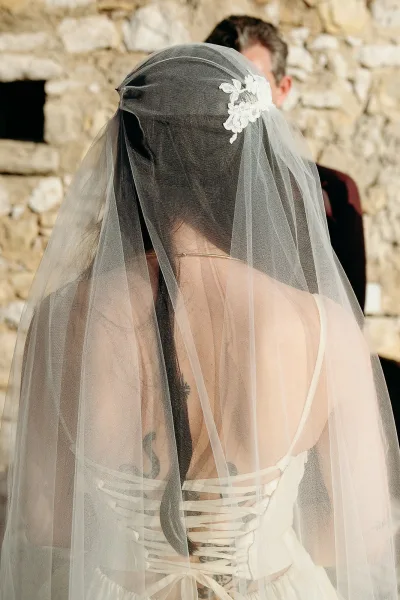 Wedding veil drapes over a strapless corset-back gown with lace applique veil details, bride tattoo visible, by a stone wall outdoors