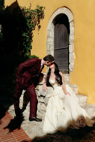 Wedding kiss portrait of bride and groom kissing on stone steps by an arched wooden door, sunlight on a yellow stucco wall and vines