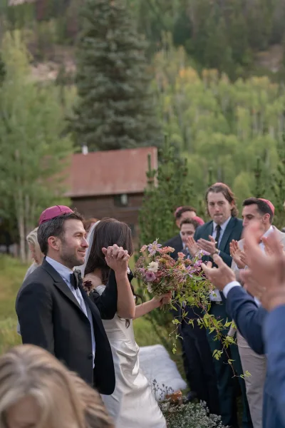 Recessional moment as bride and groom walk up the aisle, bride holding bouquet and veil flowing, guests clap by rustic cabin in mountains