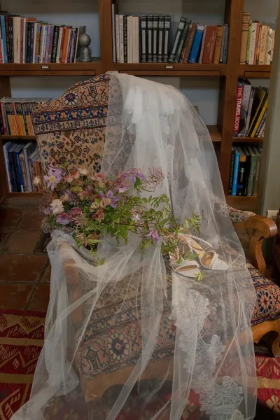 Bridal accessories with a lace wedding veil draped over a chair beside pearl heels and a wildflower bouquet in a book-lined room