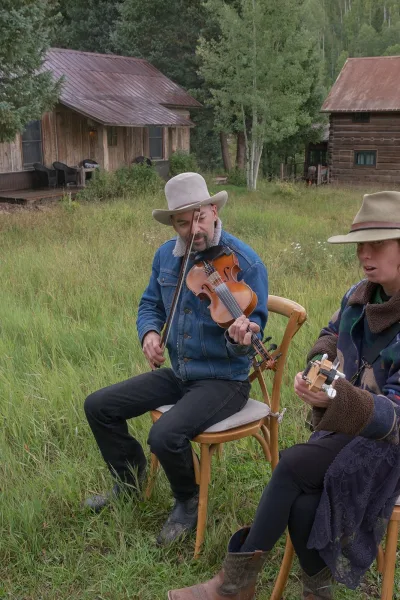 Wedding musicians play live wedding music with a violin accent and guitar, seated in a grassy field near rustic cabins and trees