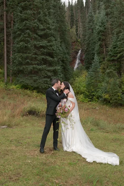 Wedding kiss portrait of bride and groom kissing, her long veil flowing with bouquet in hand, beside a waterfall in evergreen forest meadow
