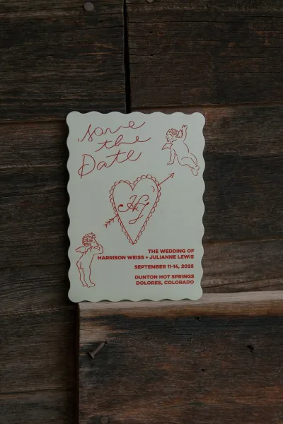 Save the date card with red ink typography and a scalloped edge, featuring cupid and heart illustrations, resting on rustic wood boards