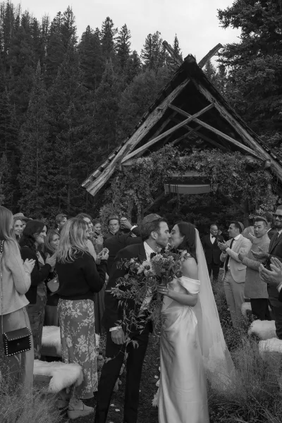 Ceremony kiss as bride in off the shoulder dress and veil holds bouquet, groom in tux, guests cheer under wooden arch in forest setting