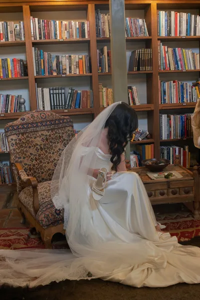 Bridal portrait of a bride looking down in a patterned armchair, lace veil and satin dress draped in a moody library of bookshelves