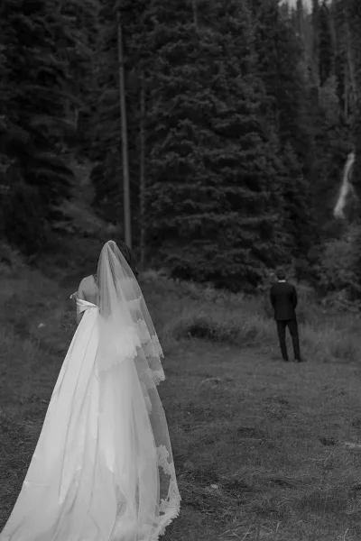 First look moment as bride approaches groom from behind, lace-edged veil trailing over grass in a forest clearing, bouquet in hand