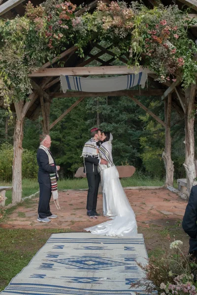 Wedding kiss under a floral chuppah as bride in gown and groom in tuxedo and kippah embrace on a forest lawn patio