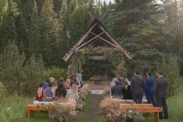 Ceremony setup with wooden ceremony arch draped in floral garland, benches and rugs lining the aisle in a sunlit pine forest.