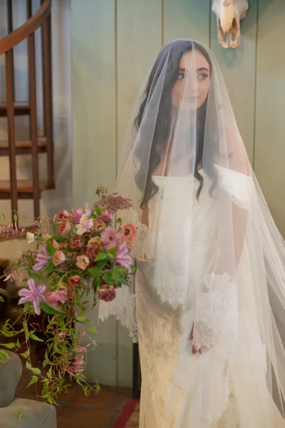 Bridal portrait of a bride with veil over her face holding a pink bouquet with greenery beside a wood spiral staircase and green wall indoors