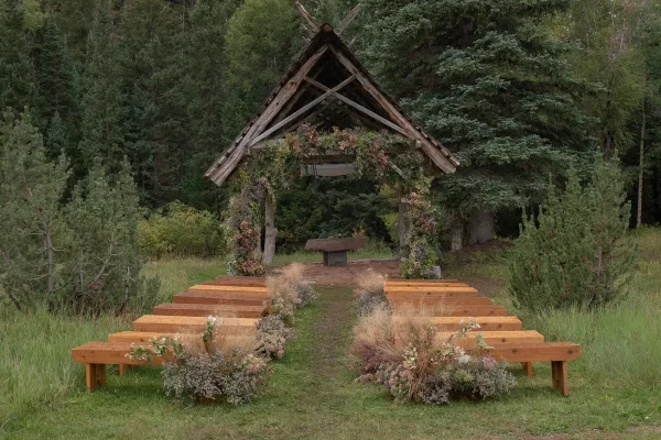 Ceremony setup with outdoor ceremony seating on wood benches facing a wooden arbor with floral garland, aisle florals, and dried grasses in a forest clearing