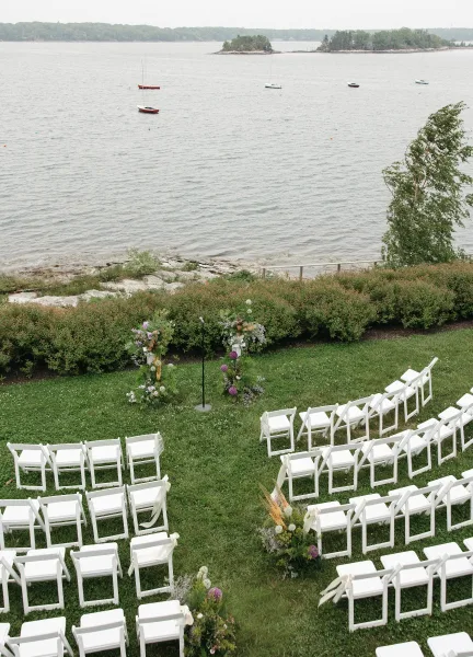 Ceremony setup for a waterfront ceremony with white folding chairs on a grass lawn, sailboats gliding beyond a rocky shore and hedges.
