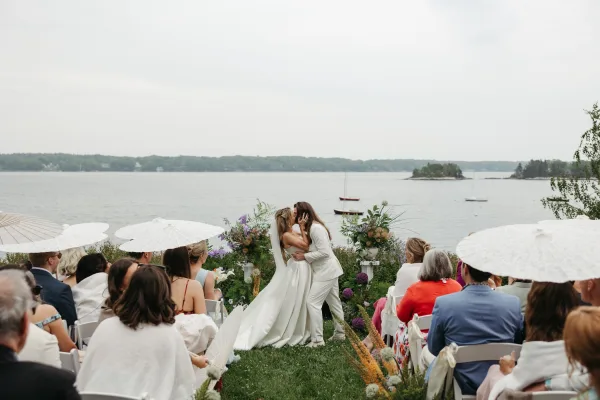 Wedding ceremony kiss at a waterfront wedding ceremony as a bride embraces her partner in a white suit, with sailboats on the distant shore.