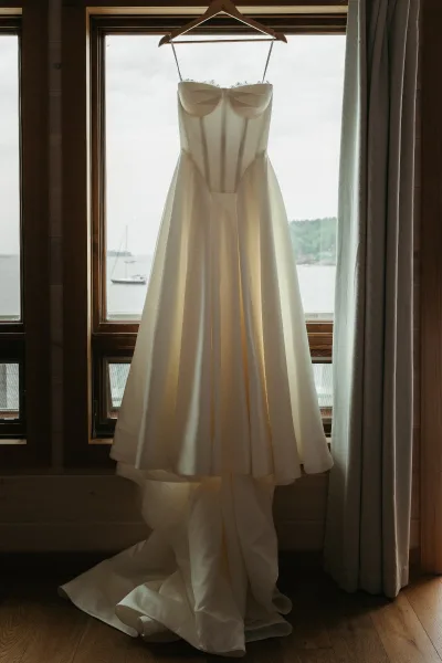 Wedding dress, an A-line wedding dress on a hanger before a window, overlooking a lake with a single sailboat, trees, and sky.
