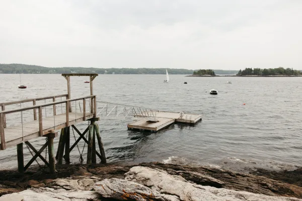 Waterfront dock and wooden jetty leading to a moored sailboat, with small islands and a distant tree line under an overcast sky.
