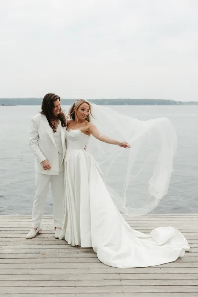 Couple portrait of newlyweds by the water on a wood dock, bride lifting her veil beside a calm lake under an overcast sky.
