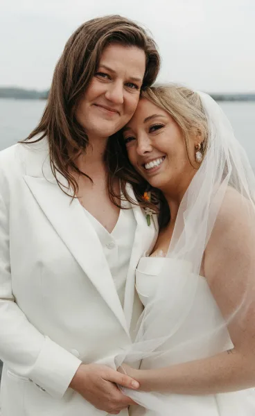 Couple portrait of two brides cheek to cheek by the water, one in a white suit, sharing warm wedding day smiles.