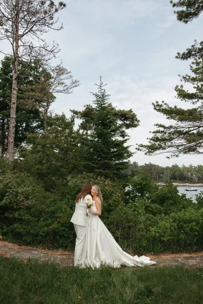 Couple portrait of two brides standing on a stone path by a lakeside with evergreens, one holding a small orange and white bouquet.