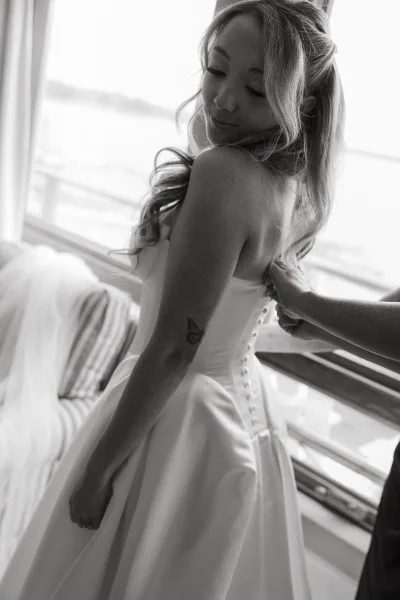 Bride getting ready, buttoning her wedding dress with covered buttons by a bright window overlooking the water.