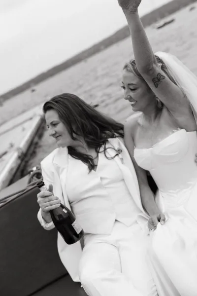 Wedding celebration moment with a wedding couple on a boat popping a champagne bottle, with water, docked boats, and open sky behind them.