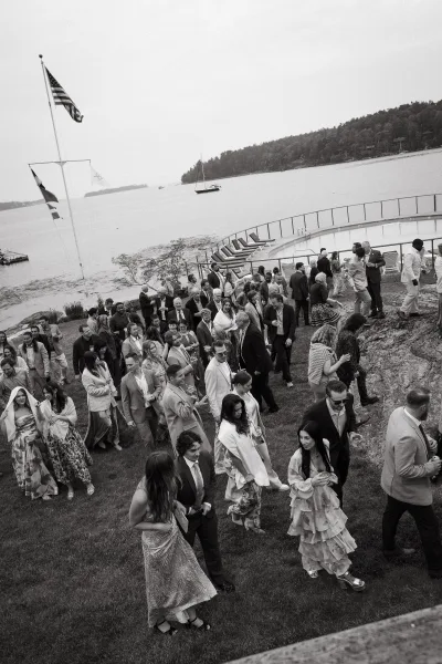 Cocktail hour with wedding guests mingling, shawls catching the breeze by a waterfront pool as sailboats dot the cloudy horizon.