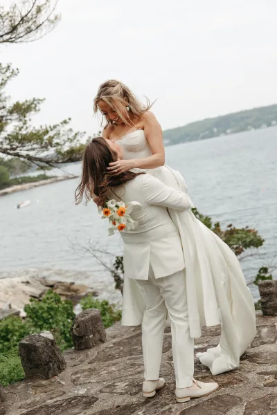 Couple portrait in a wedding lift pose by a lakeside rocky shoreline, partner lifting the bride holding calla lilies.