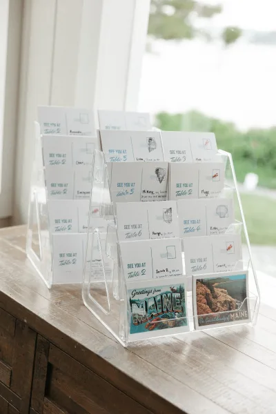 Escort card display featuring wedding seating cards in an acrylic holder, arranged on a wood table by a window with greenery and water view.