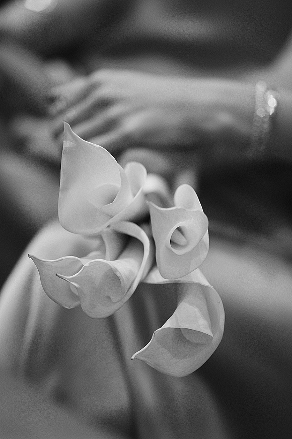 Calla lily bouquet of white blooms with ribbon wrap and bracelet detail, held close-up against blurred hands and soft bokeh background