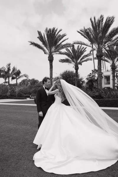 couple portrait of a bride and groom outdoors under palm trees, her cathedral veil trailing as they pose on a sunlit lawn near a stately building