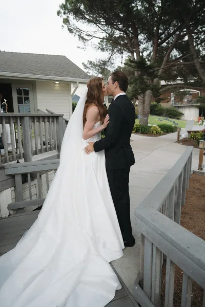 Wedding kiss portrait of newlyweds on a wooden deck, an outdoor wedding portrait framed by railings and trees, her veil trailing to the garden path.