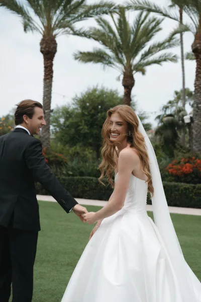 Couple portrait of a bride and groom holding hands on a sunlit lawn beneath palm trees, her veil catching light as garden string lights glow.