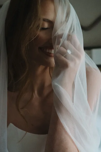 Bridal portrait of a bride under veil, smiling softly as she lifts the tulle to reveal a sparkling diamond ring and a simple, elegant pose.